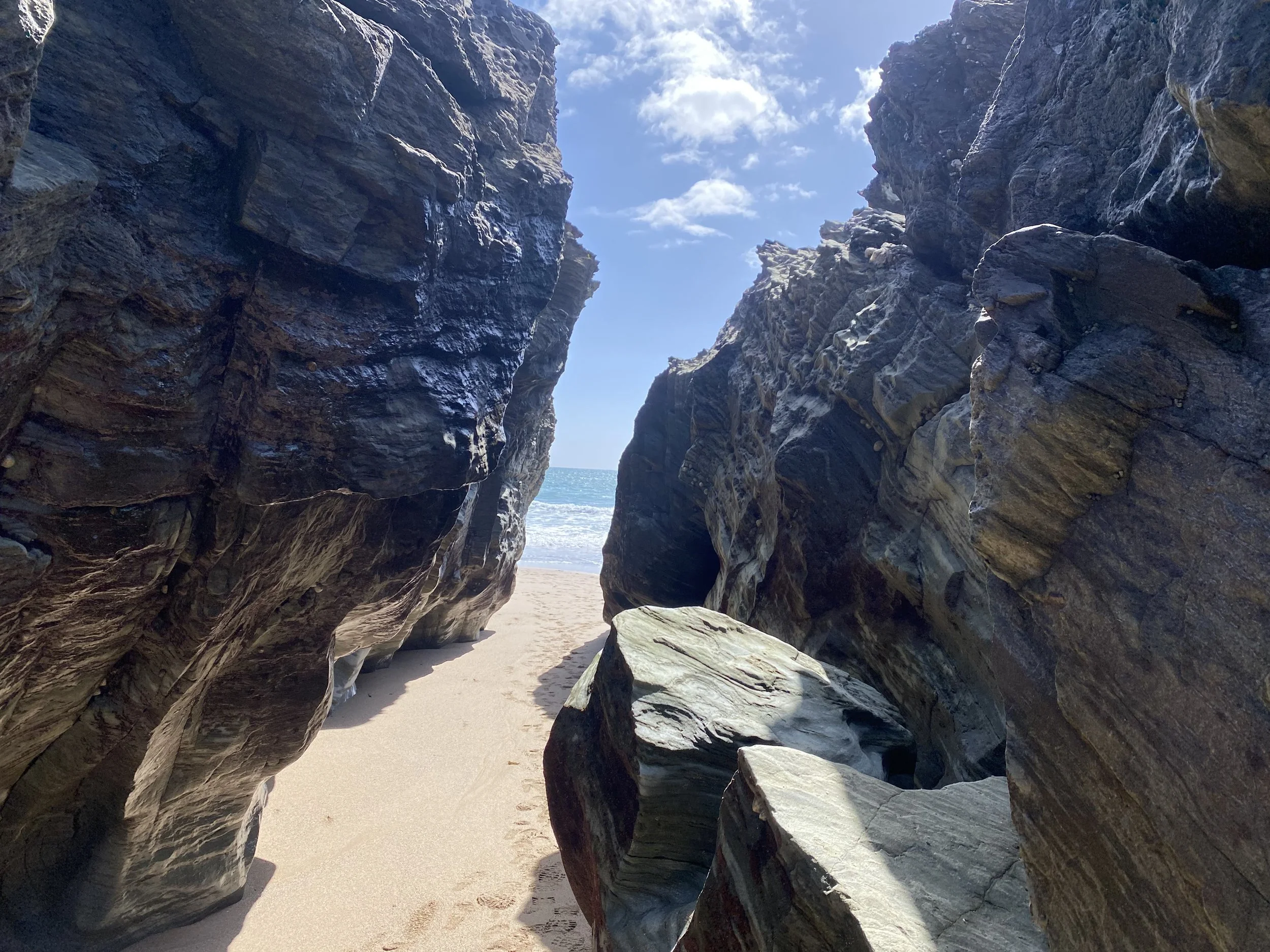 View of a sandy beach through a narrow gap between large, dark rocks. The sky is partly cloudy and the ocean can be seen in the background.