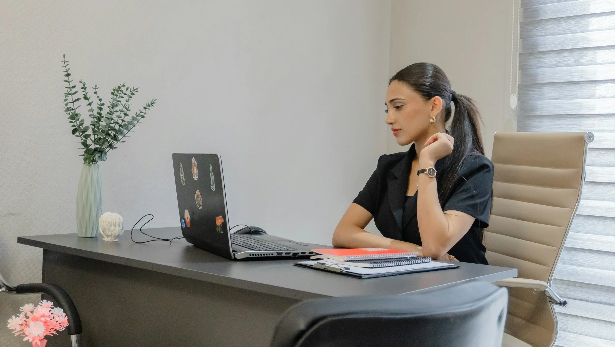 A woman with dark hair tied in a ponytail, wearing a black top, is sitting at a gray office desk, looking at her laptop. The desk has some notebooks, a pen, and a red folder. There is a vase with green foliage and a small decorative object on the desk, and a window with striped blinds behind her.