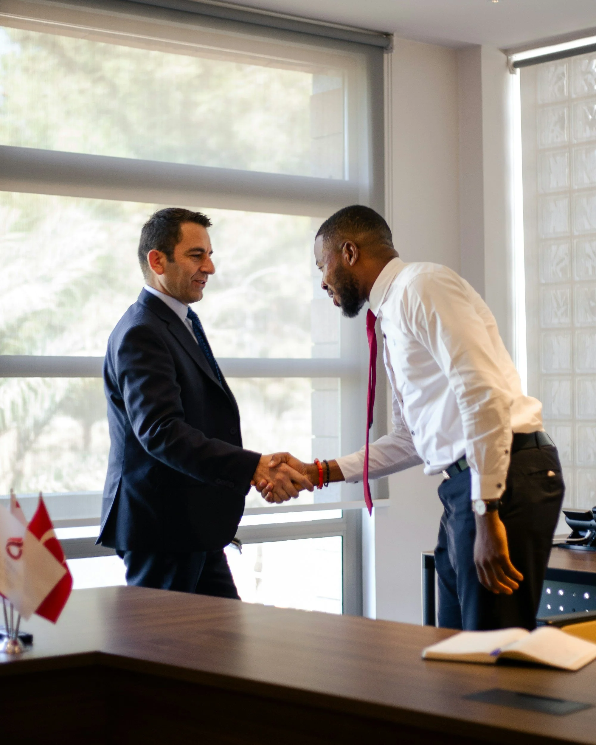 Two men in business attire shaking hands in an office with large windows and flags on the desk.