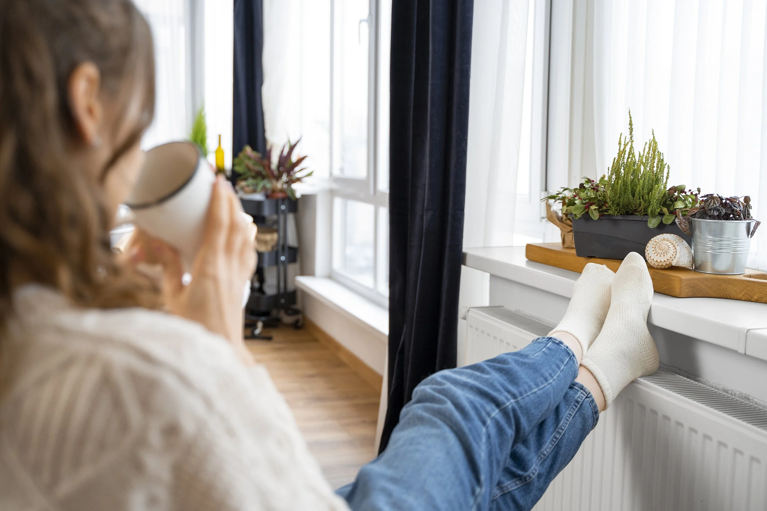 Person with long hair drinking coffee or tea while relaxing with feet up on a white windowsill, surrounded by potted plants and sunlight filtering through curtains.