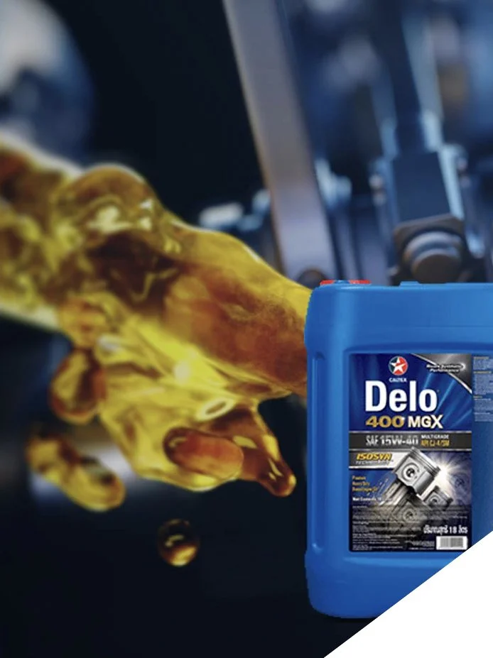 A blue container of motor oil, labeled Delo 400 MGX, is placed next to a vehicle engine with yellowish oil pouring into or inflating out of the engine.