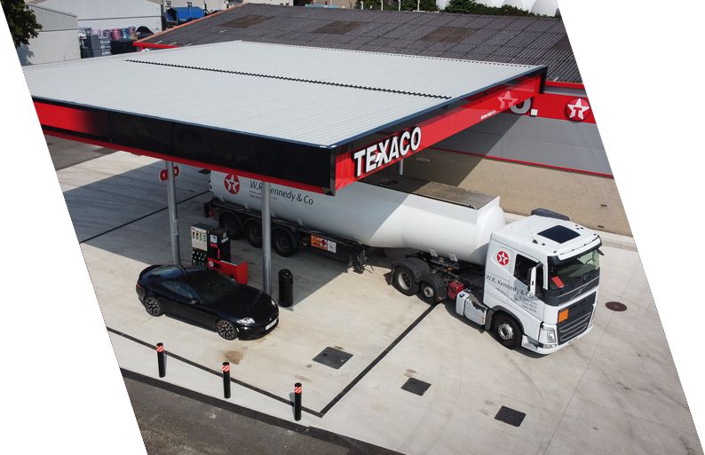 A Texaco gas station with a tanker truck refueling a black car, and a white truck parked nearby, under a canopy with the Texaco logo.