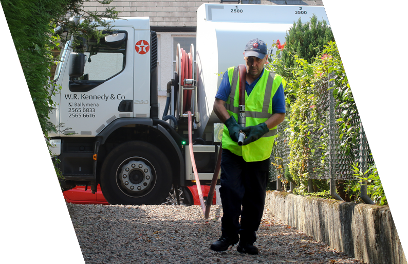 Worker in safety vest and gloves using a hose connected to a sanitation truck on a gravel pathway next to a fence with plants.