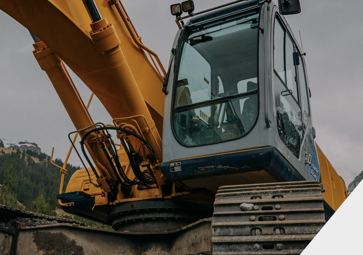 Close-up view of an excavator with a yellow arm and black tracks parked outdoors with mountains in the background