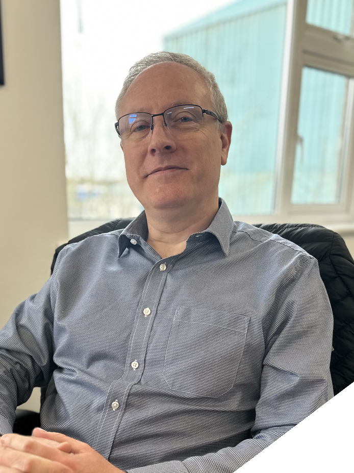 A middle-aged man with glasses sitting in an office chair near a window, wearing a blue collared shirt, looking at the camera.