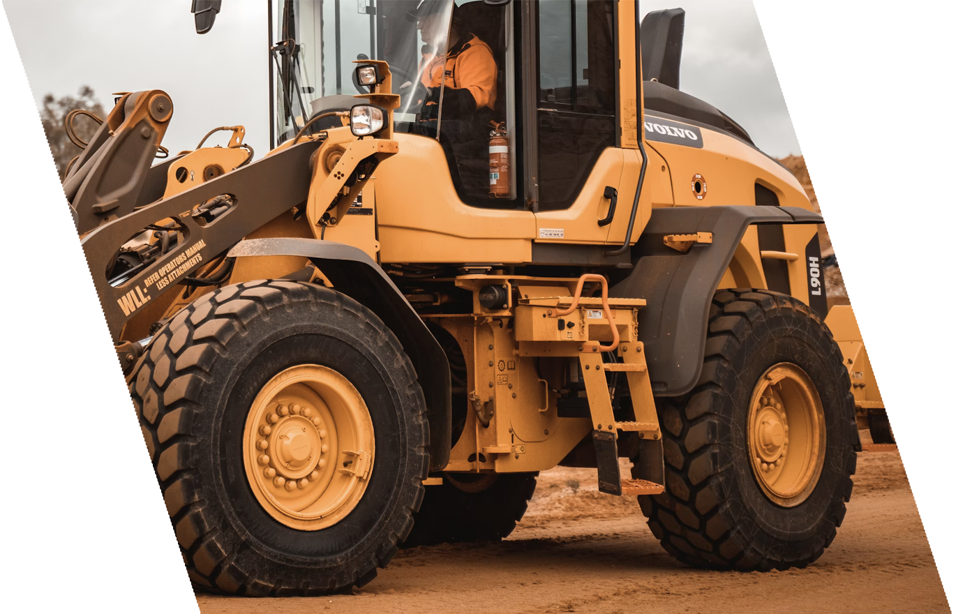A yellow construction bulldozer on a dirt surface with a person in the cab, wearing an orange jacket, and a fire extinguisher mounted inside the cab. The bulldozer is marked with the brand 'Volvo' and has large, rugged tires.