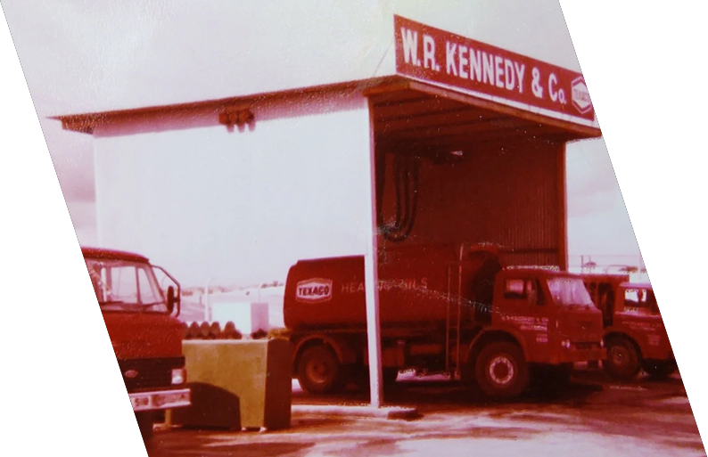 Vintage gas station with a large red sign reading 'W. R. Kennedy & Co.' and a Texaco fuel truck parked under the roof.
