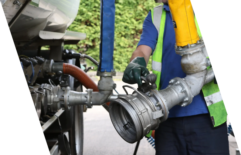 A worker in a safety vest and gloves holding a large industrial pipe component near a vehicle or equipment outdoors, with green trees in the background.