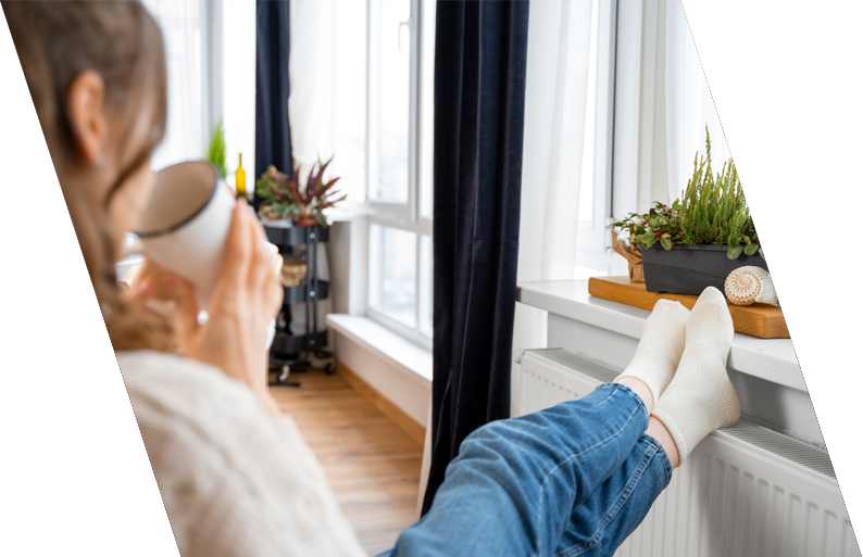 Person sitting on a window sill, drinking from a mug, with their legs crossed, overlooking a bright living room with potted plants and hardwood flooring.
