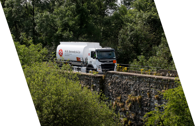 A white tanker truck labeled W.R. Kennedy & Co driving on a narrow road surrounded by green trees and foliage.