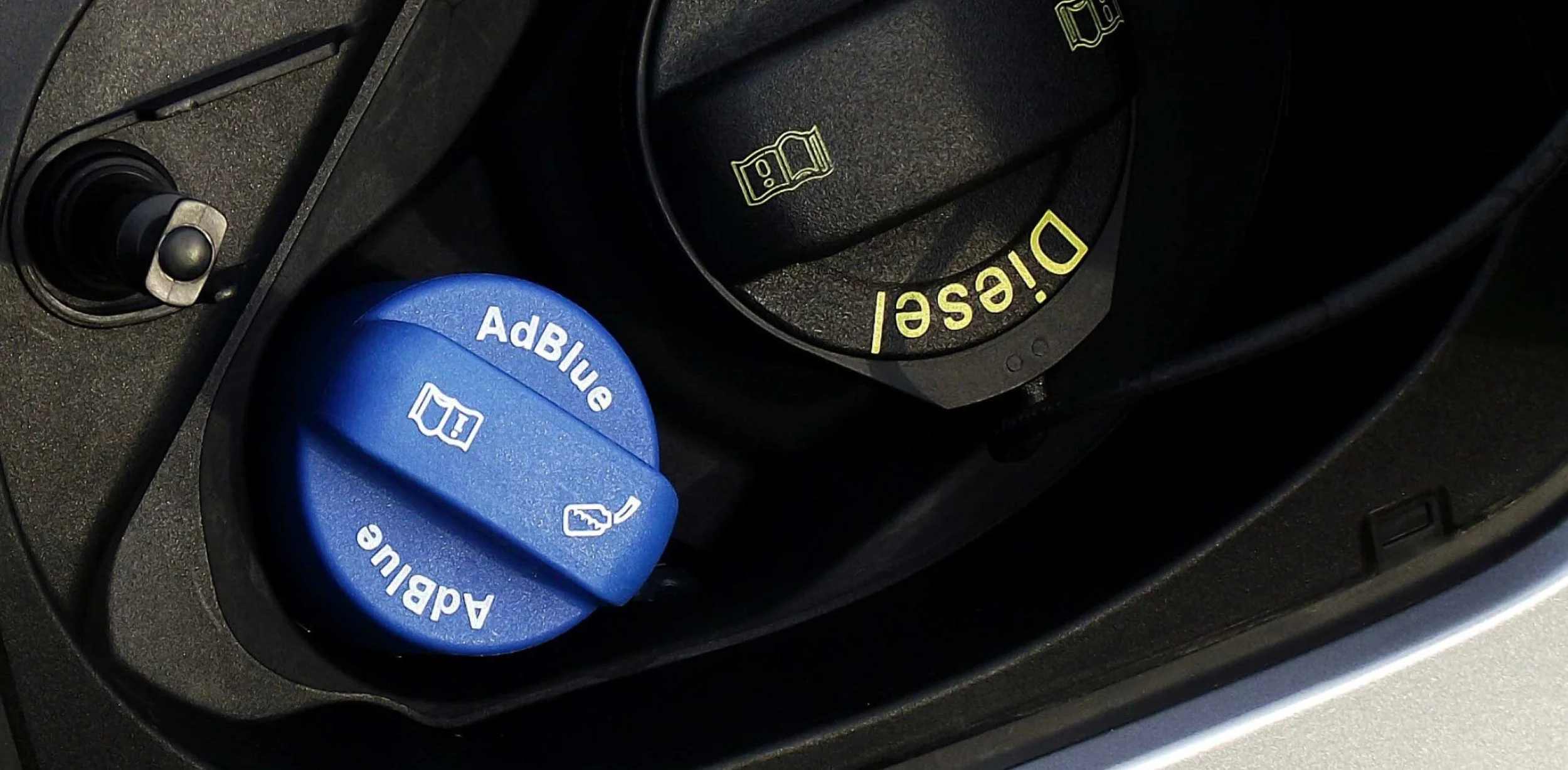 A close-up of a car's dipstick and oil cap. The yellow cap is labeled 'DIPSE' and the blue cap is labeled 'AdBlue' with an icon of a fuel pump.