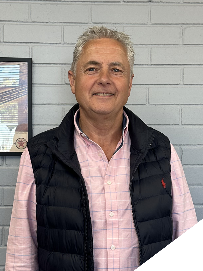 A middle-aged man with gray hair wearing a pink checkered shirt and a black puffer vest standing in front of a white brick wall.