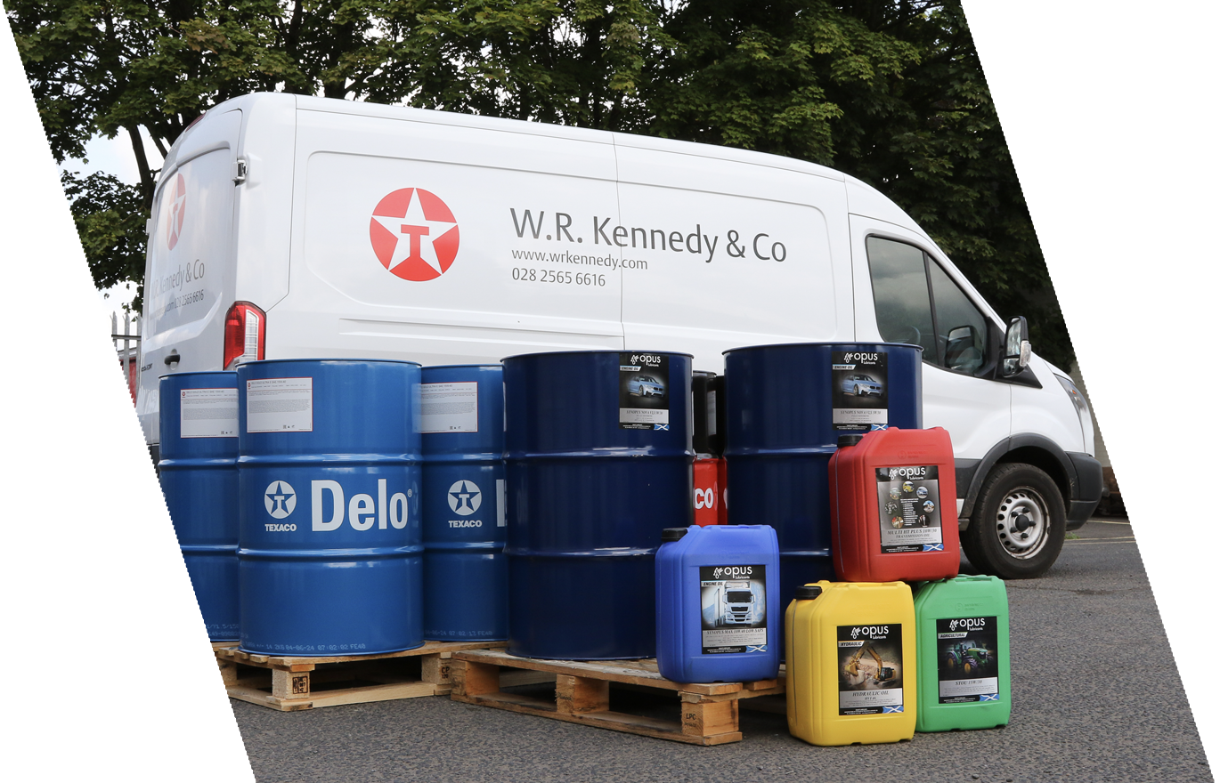 A white W.R. Kennedy & Co van parked outdoors with assorted oil drums and containers in front, including blue, red, yellow, and green.