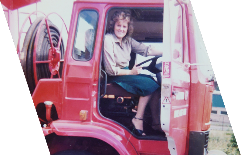 A woman sitting in the driver's seat of a red utility truck, holding the steering wheel and smiling at the camera.