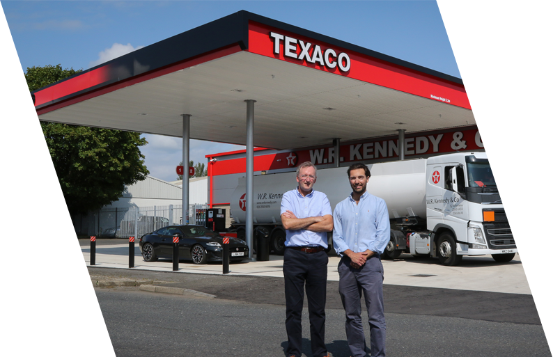 Two men standing under a Texaco gas station canopy with a tanker truck and cars in the background.