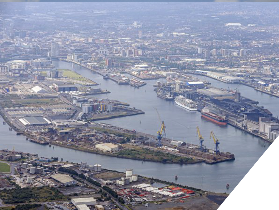Aerial view of a busy industrial port with cranes, ships, and various warehouses along a river in an urban area.