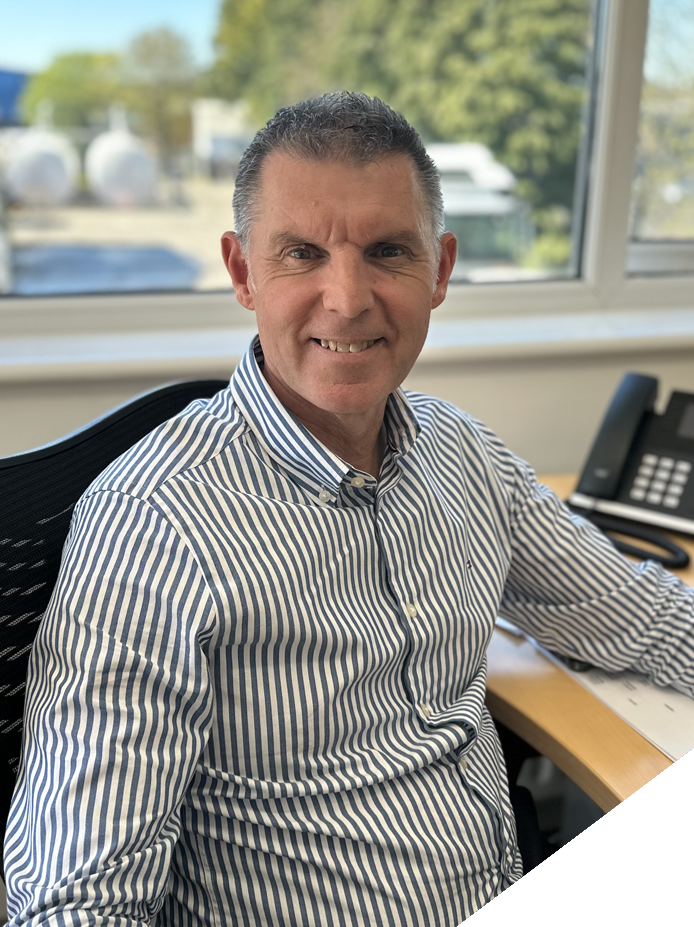 A middle-aged man with short gray hair and blue eyes sitting at a desk in an office. He is wearing a white and blue striped dress shirt and smiling at the camera. Behind him there is a large window with a view of trees and a building outside, and on the desk there is a black telephone.