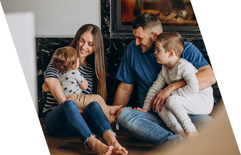 A family sitting on the floor near a fireplace, smiling and playing together. The mother is holding a young child, and the father is with an older child, all engaging in a joyful moment.