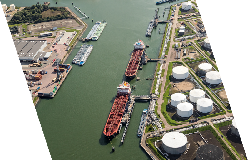 Aerial view of an industrial port with three large cargo ships docked, surrounded by storage tanks, pipelines, and industrial buildings.