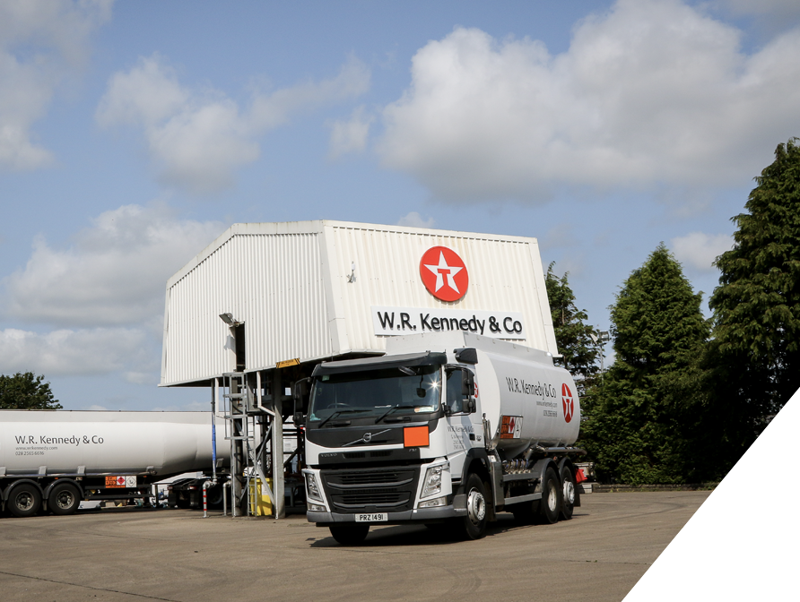 A large tanker truck with a white cab and a cylindrical tank, parked in front of a building with a sign that reads 'W.R. Kennedy & Co' and a red logo, under a partly cloudy sky, surrounded by green trees.