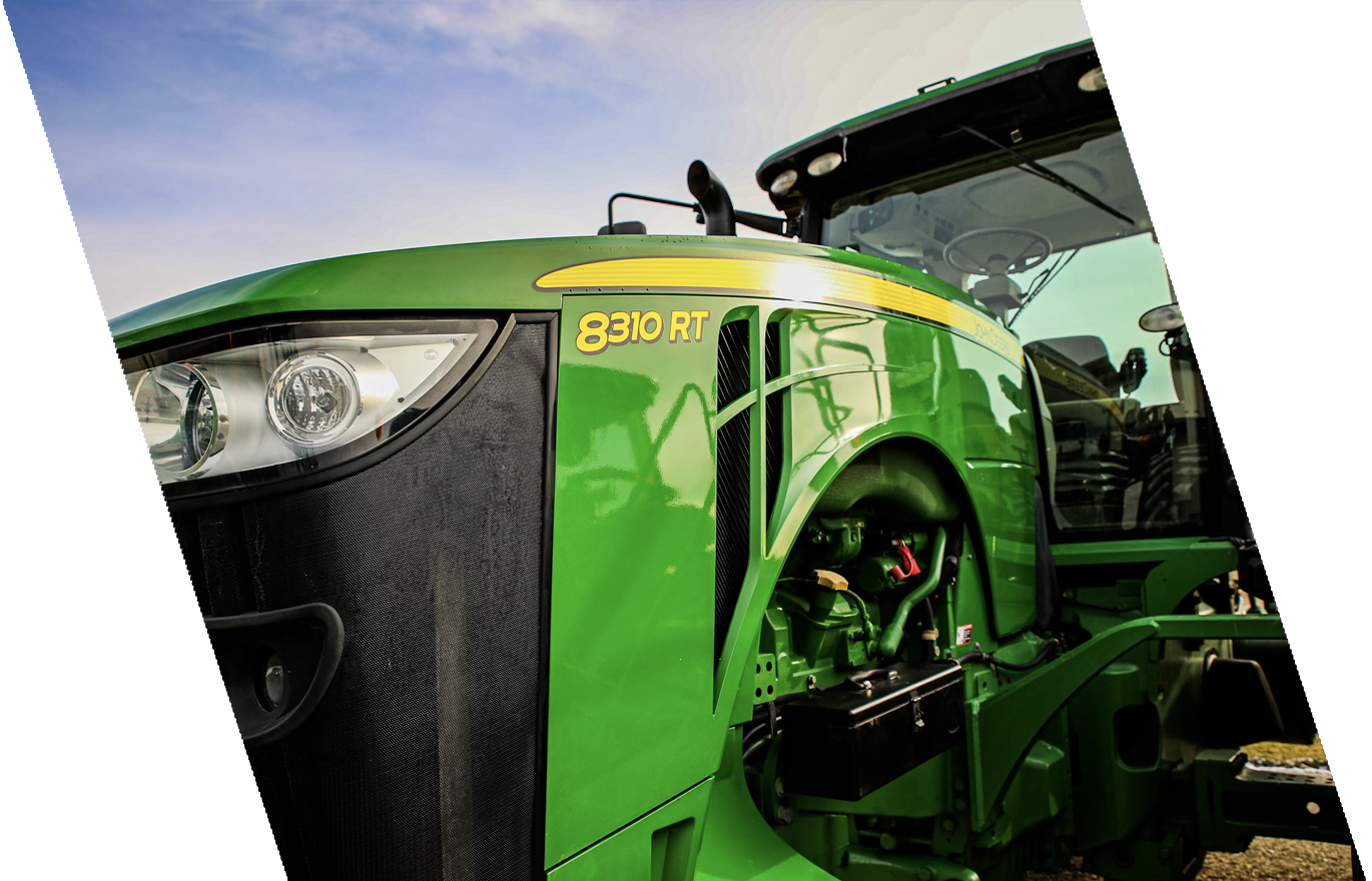 Close-up of a green John Deere 8310 RT tractor showing the front headlight, side panel, and part of the cockpit with a clear sky in the background.