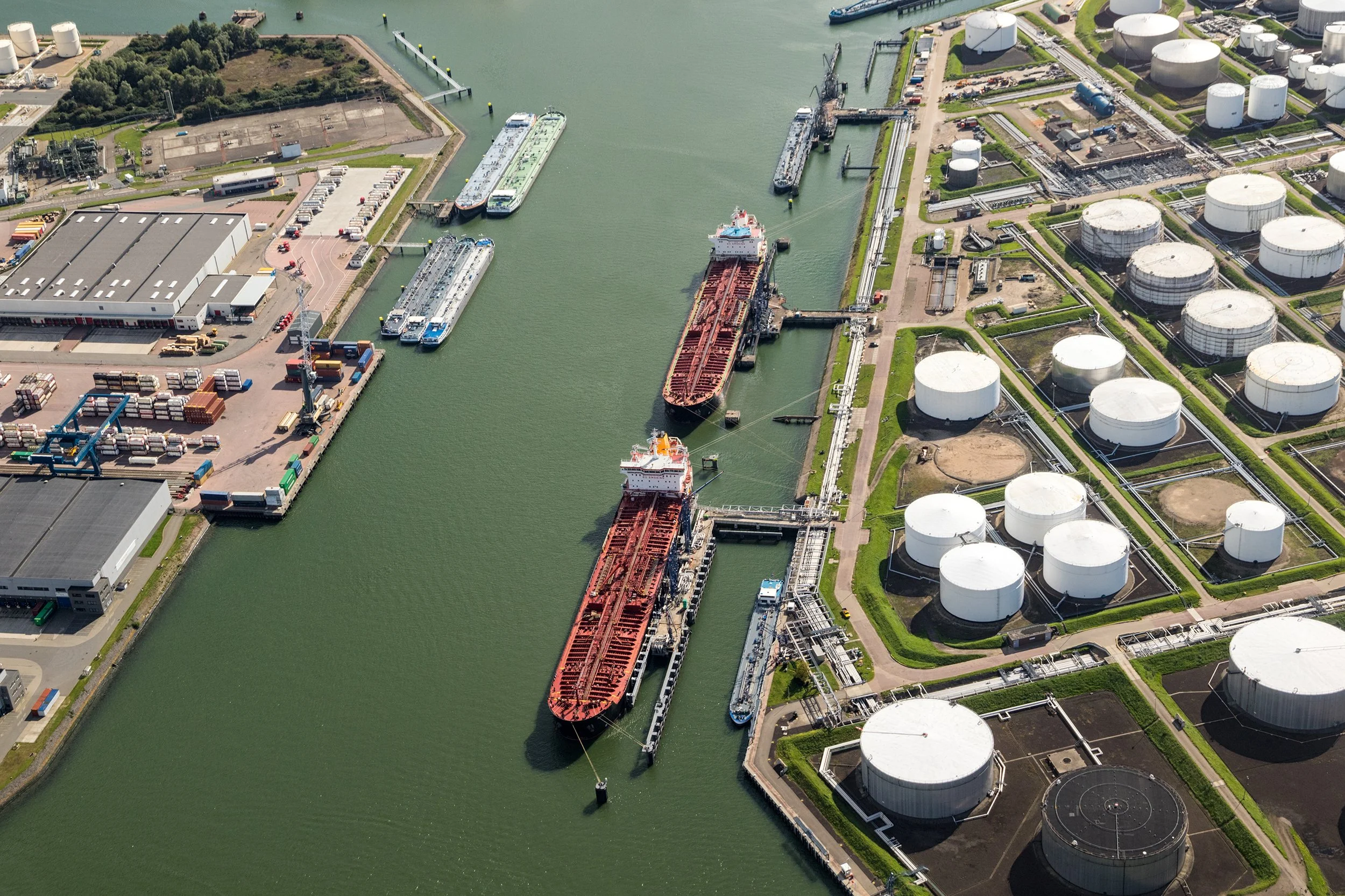 An aerial view of an industrial port with large storage tanks, two ships docked, and various buildings and containers near the water.