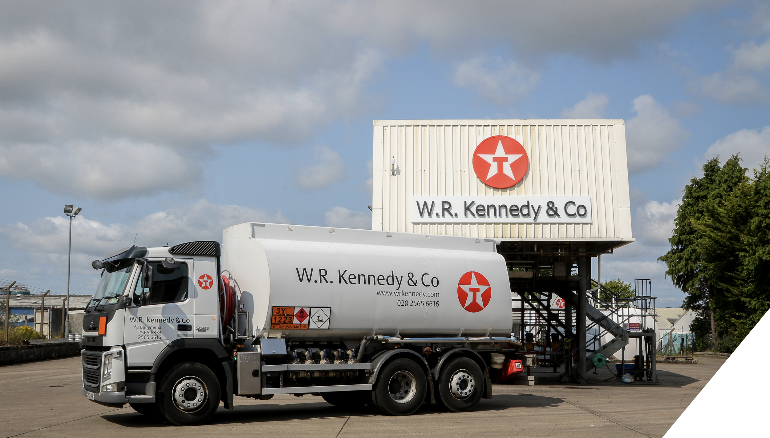 A fuel delivery truck labeled W.R. Kennedy & Co parked near a storage or service building with signage and a remote control panel, under partly cloudy skies.