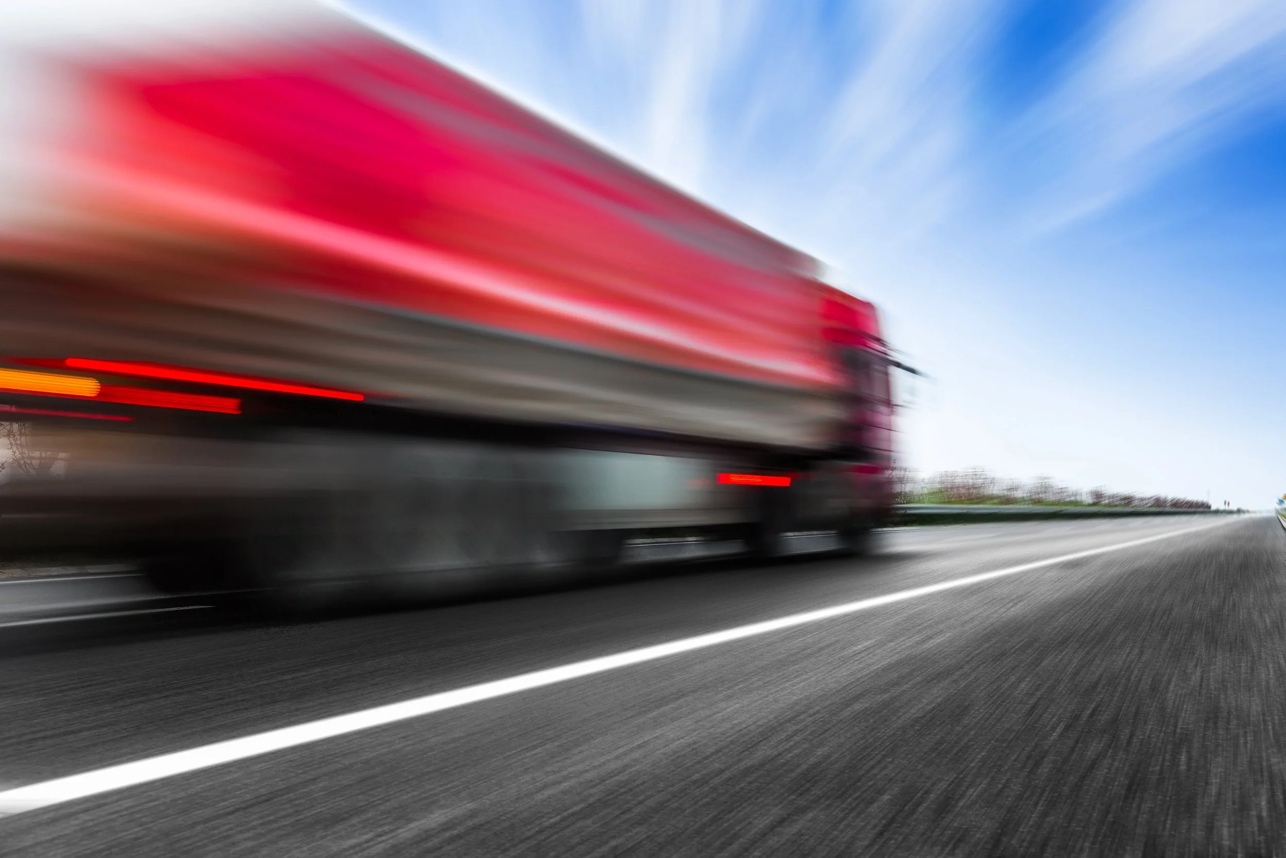 A red semi-truck driving rapidly on a highway with a blurred background and sky.