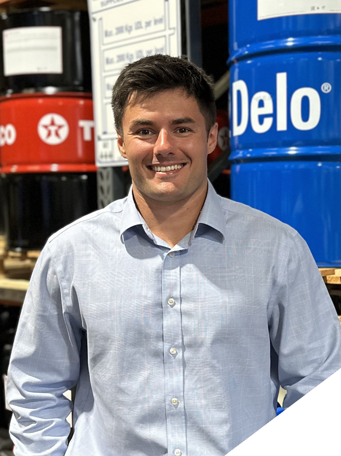 A smiling man in a light blue button-up shirt standing in a warehouse or store, with large blue and black barrels labeled "Delo" and "Texaco" behind him.