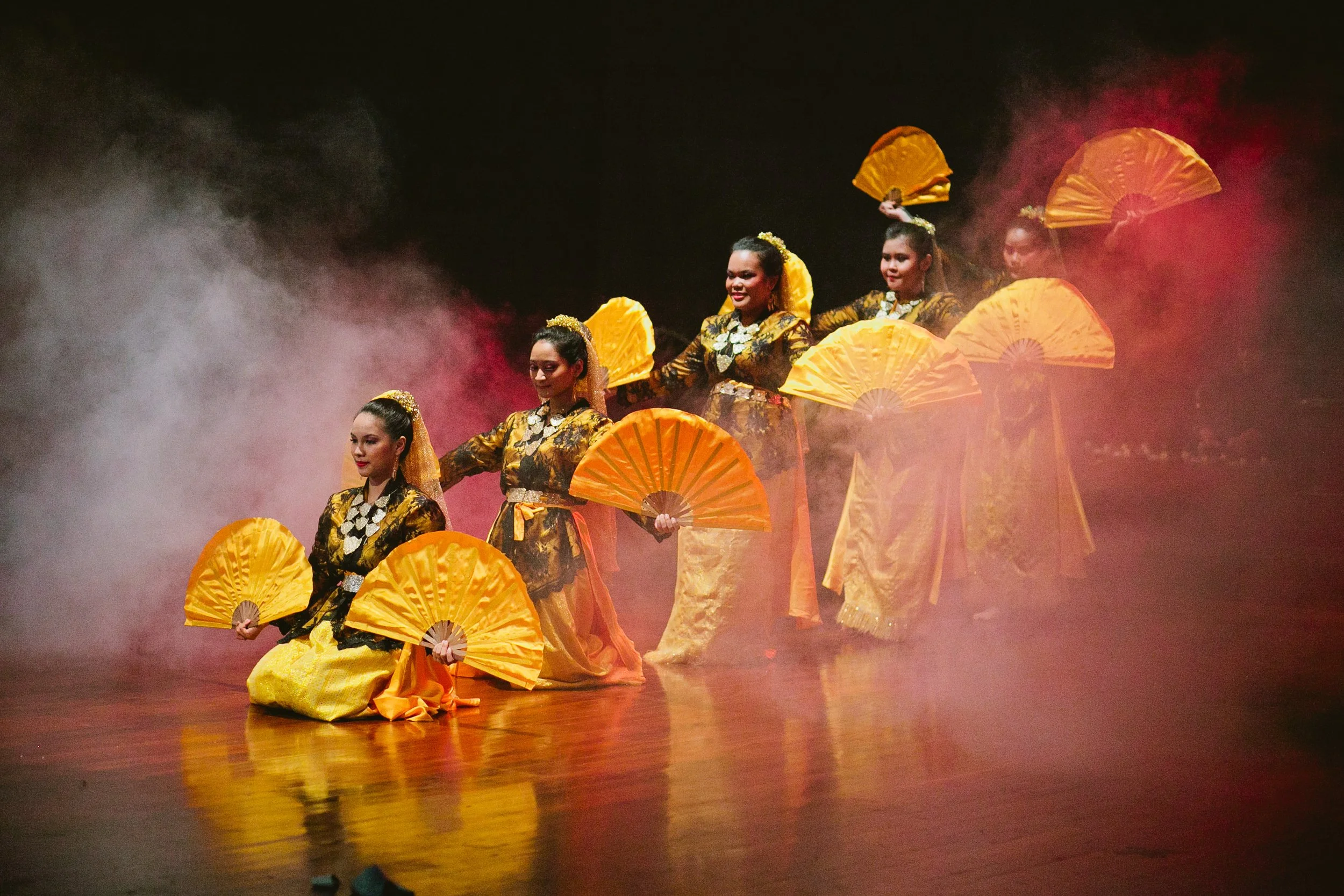 Group of women dressed in traditional Indonesian attire performing a dance with yellow fans on stage, surrounded by fog.