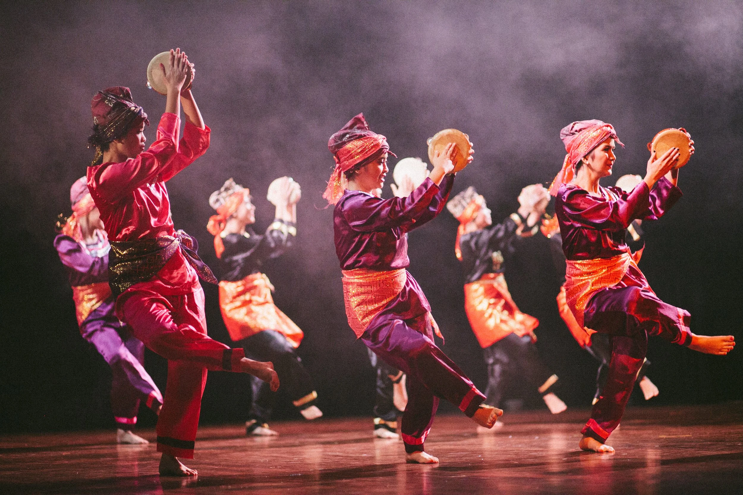 A group of performers in traditional costumes dancing on stage, holding tambourines, with a foggy background.