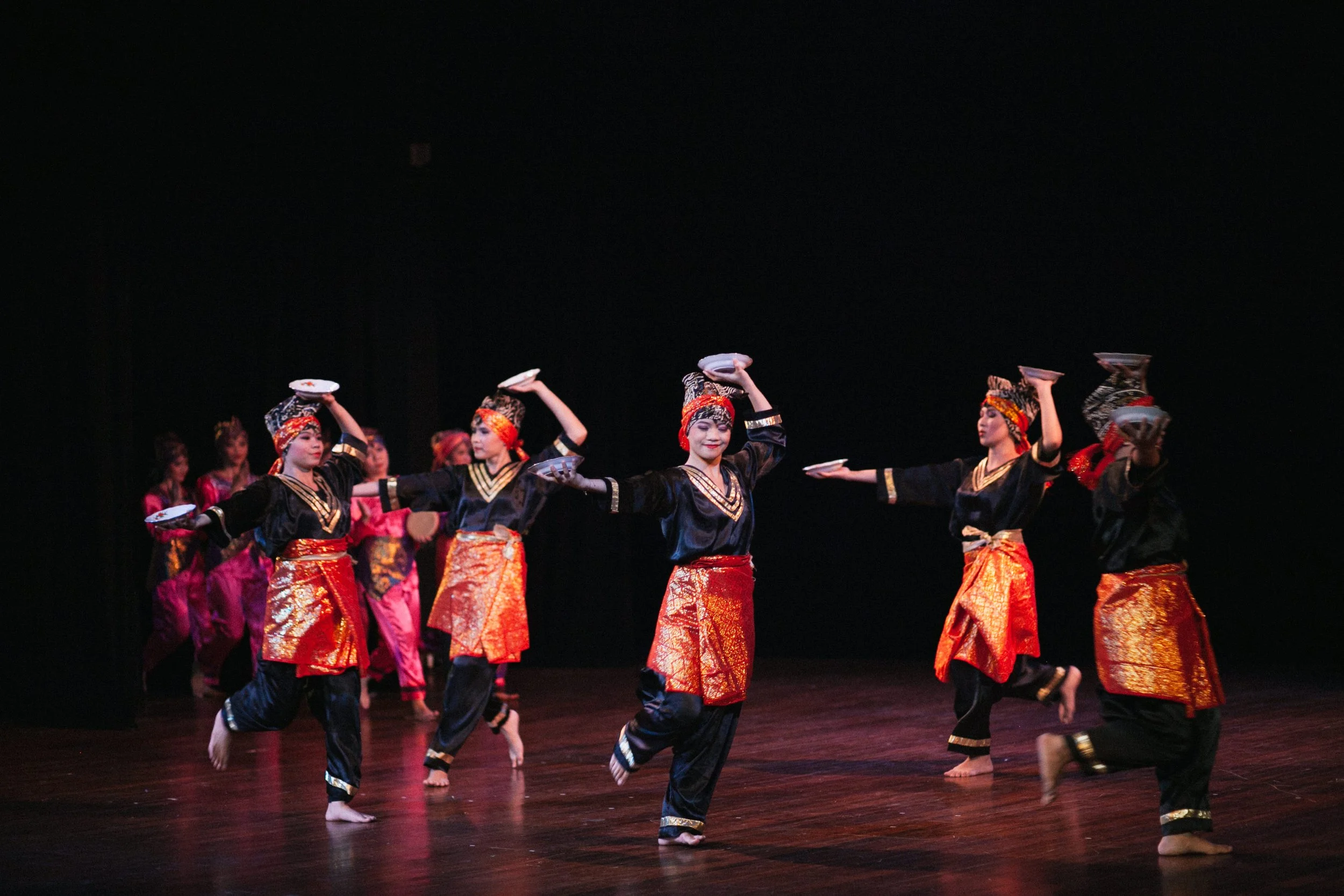 Group of dancers in traditional costumes performing on stage with black background.