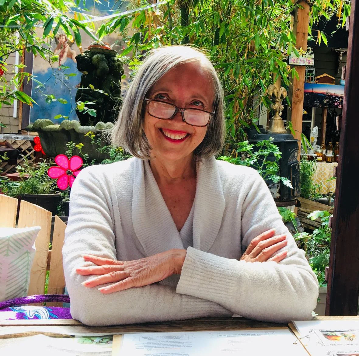 A smiling elderly woman with gray hair and glasses, sitting at a table with arms crossed in an outdoor garden setting, surrounded by lush green plants and colorful decorations.