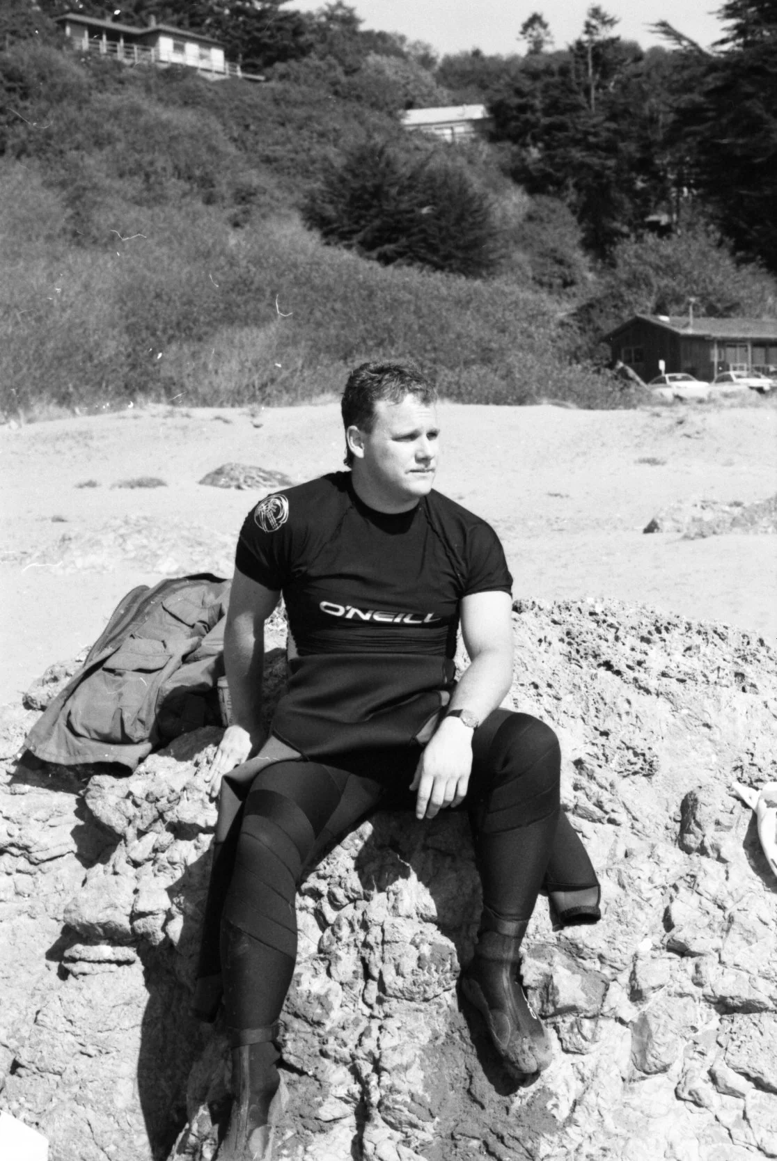 A black and white photo of a surfer sitting on a rock at the beach