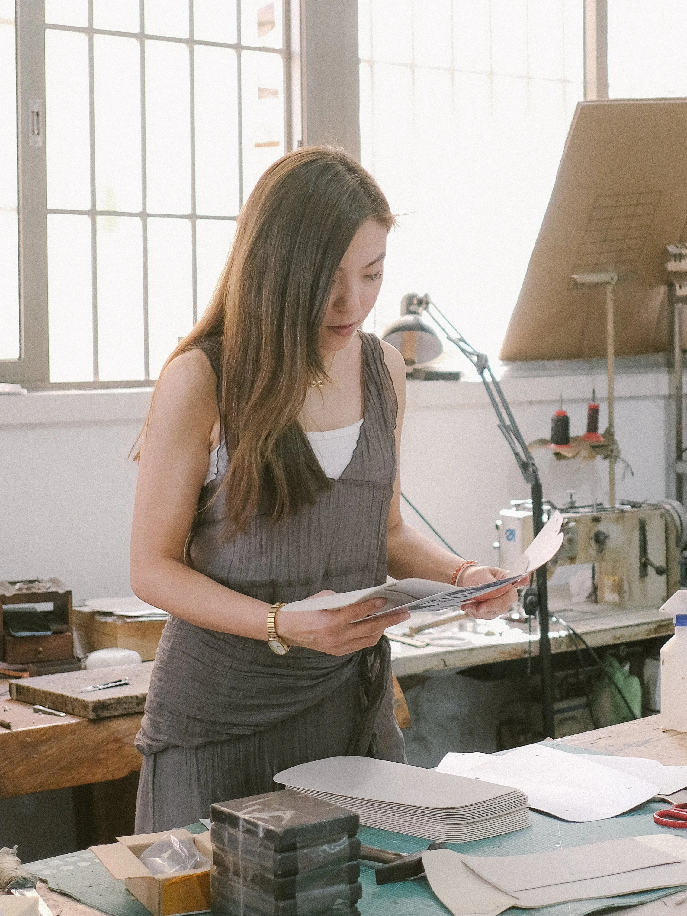 Woman with long hair looking at papers in a workspace with large windows, work tables, and tools.