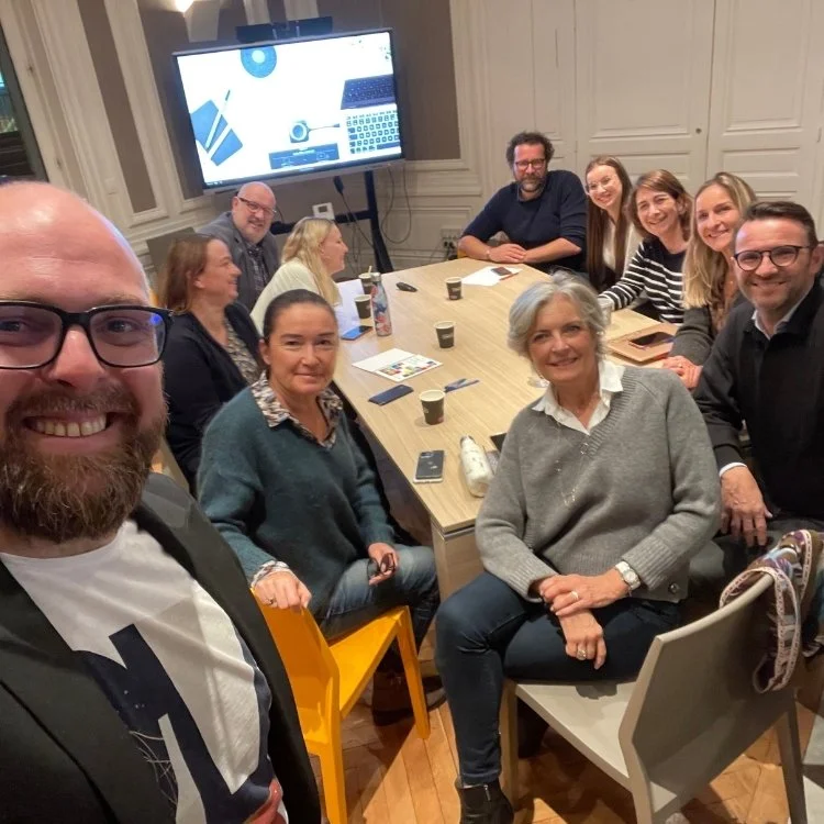 Groupe de personnes assises autour d'une table lors d'une réunion ou d'un événement social, avec un grand écran à l'arrière affichant des graphiques ou des présentations.