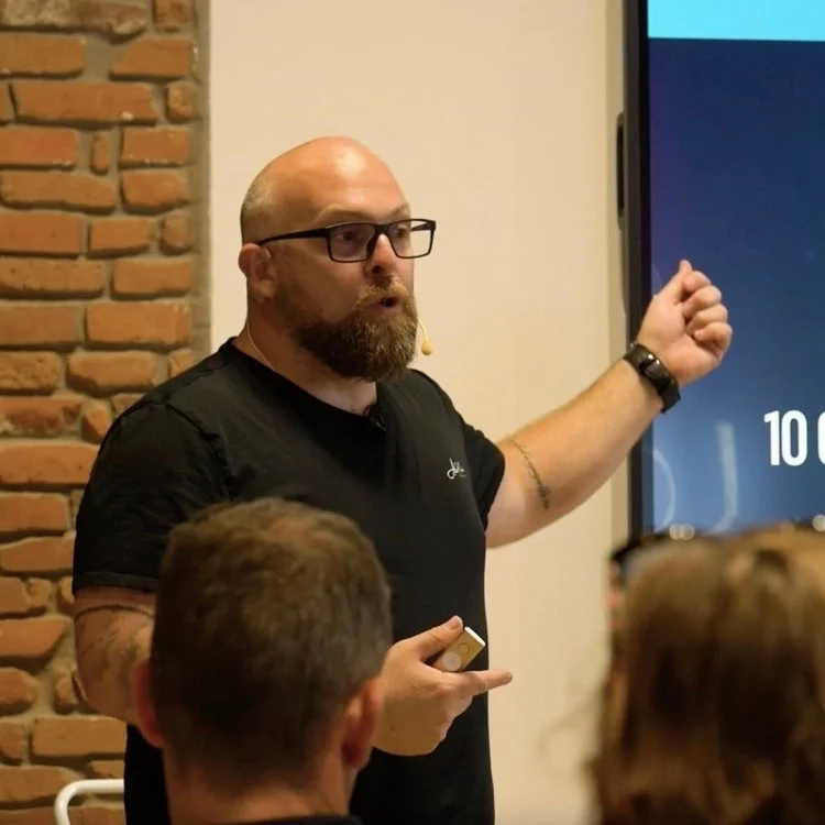 Un homme à barbe portant des lunettes et un t-shirt noir donne une présentation dans une salle avec un mur en briques en arrière-plan et un grand écran à droite.