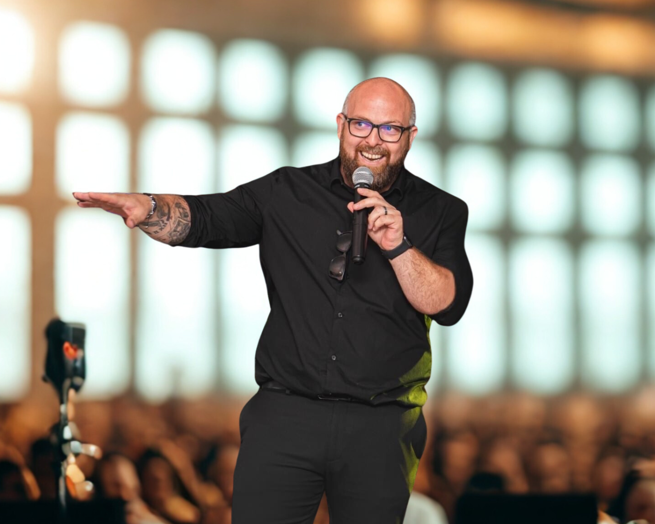 Un homme souriant avec des lunettes, tenant un microphone et faisant un geste avec la main, lors d'une présentation ou d'une conférence, dans une salle remplie de spectateurs.