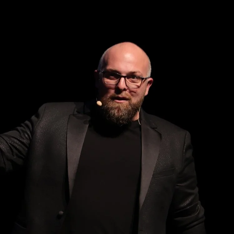 Homme avec lunettes, barbe, en costume noir, parlant lors d'une présentation ou conférence, fond noir.