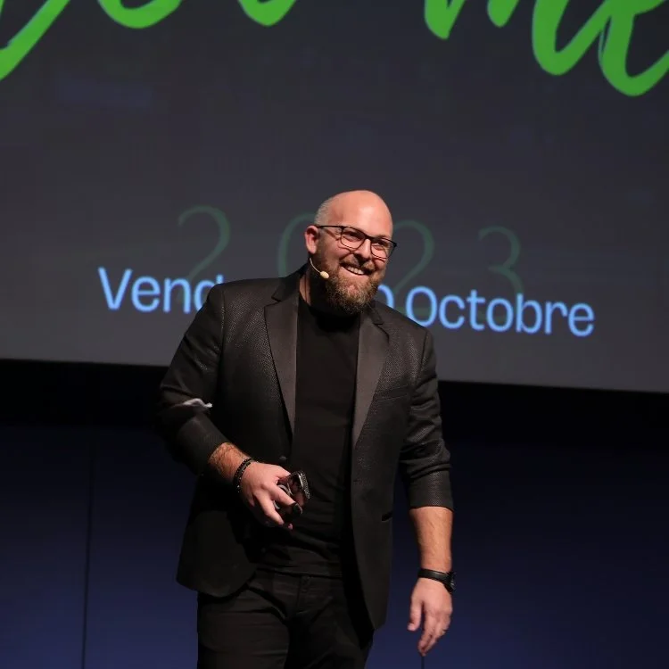 Homme souriant avec barbe et lunettes, portant une veste noire, sur scène lors d'une présentation ou conférence.