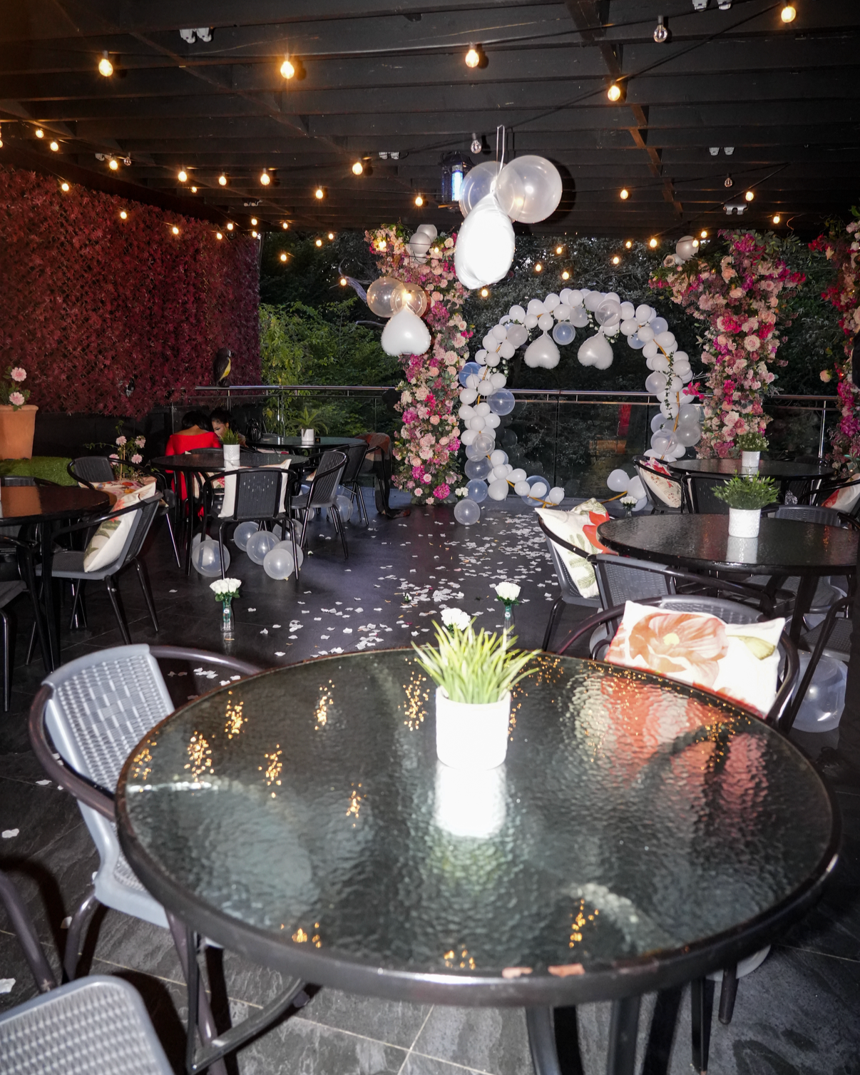 Decorated outdoor dining area with tables, chairs, floral arrangements, a balloon arch, hanging string lights, and flower petals scattered on the floor, suggestive of a celebration or event.