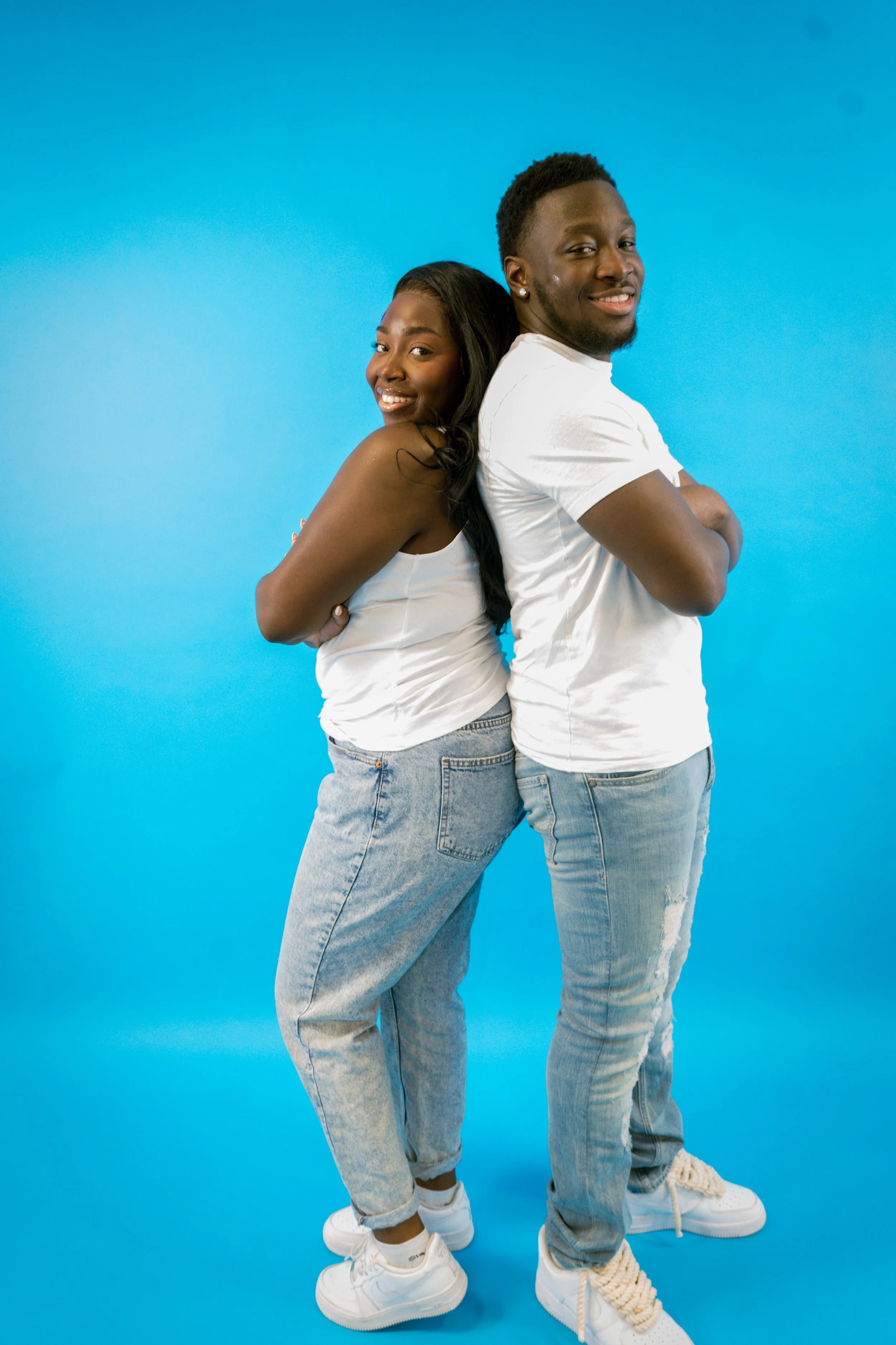 A happy Black couple standing back to back, smiling, against a bright blue background. The woman is wearing a white tank top and jeans, and the man is wearing a white T-shirt and light jeans.