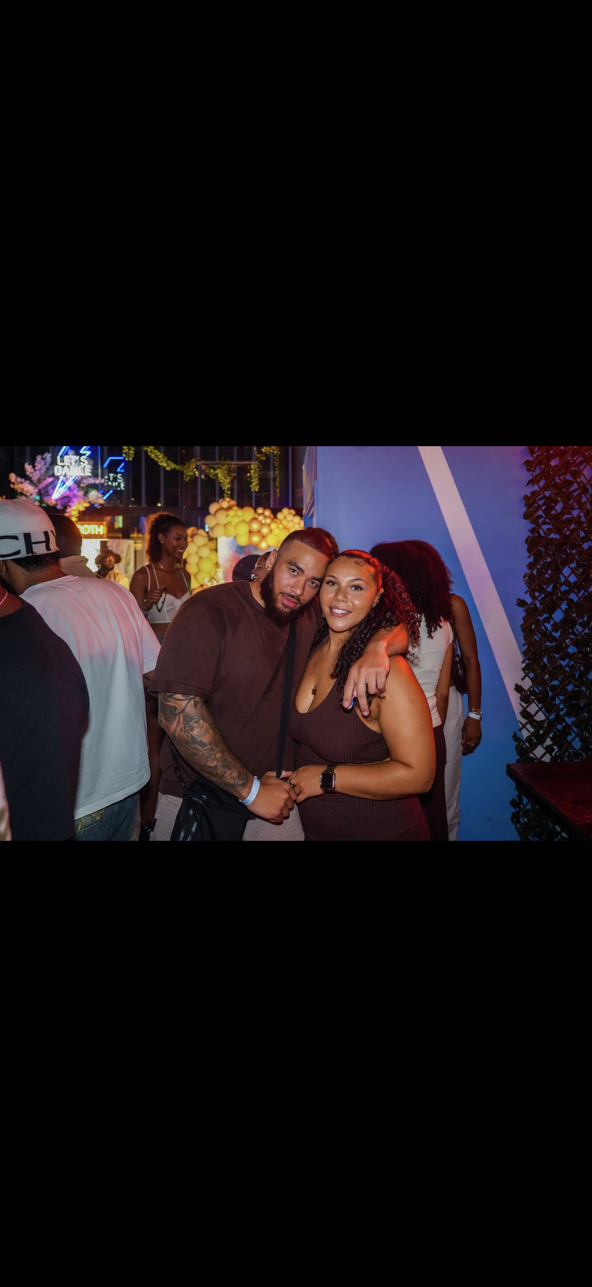 A couple posing together at a lively indoor party or club, with colorful lights and people in the background.