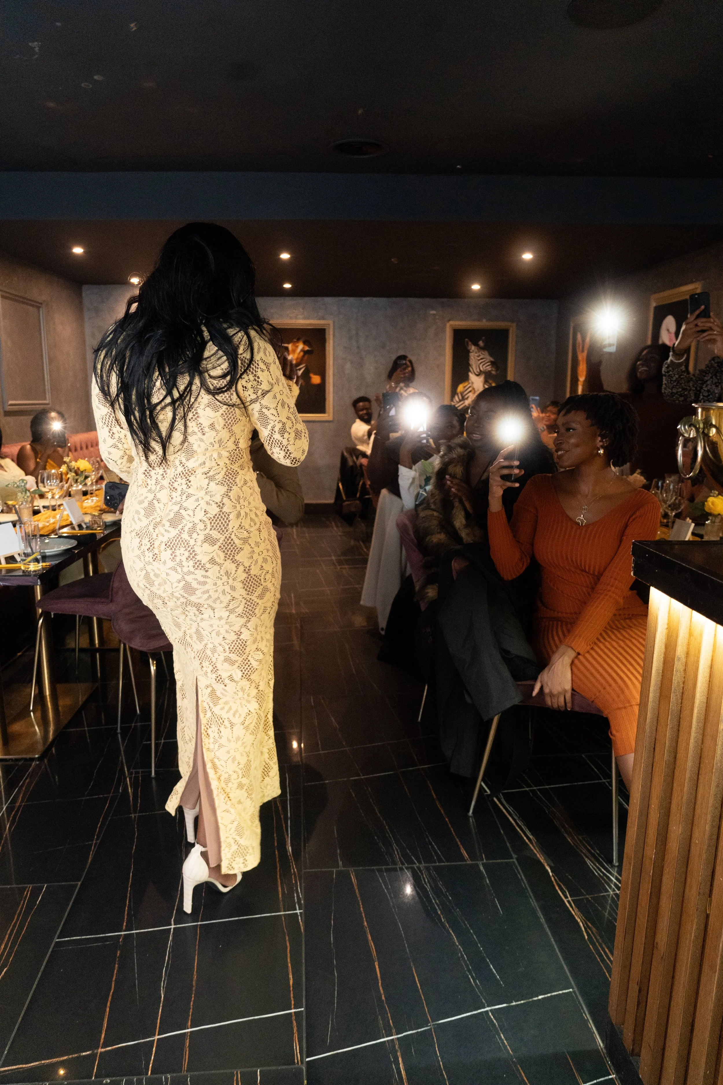 A woman in a long cream lace dress walking in a restaurant or banquet hall with people seated at tables and taking pictures of her.