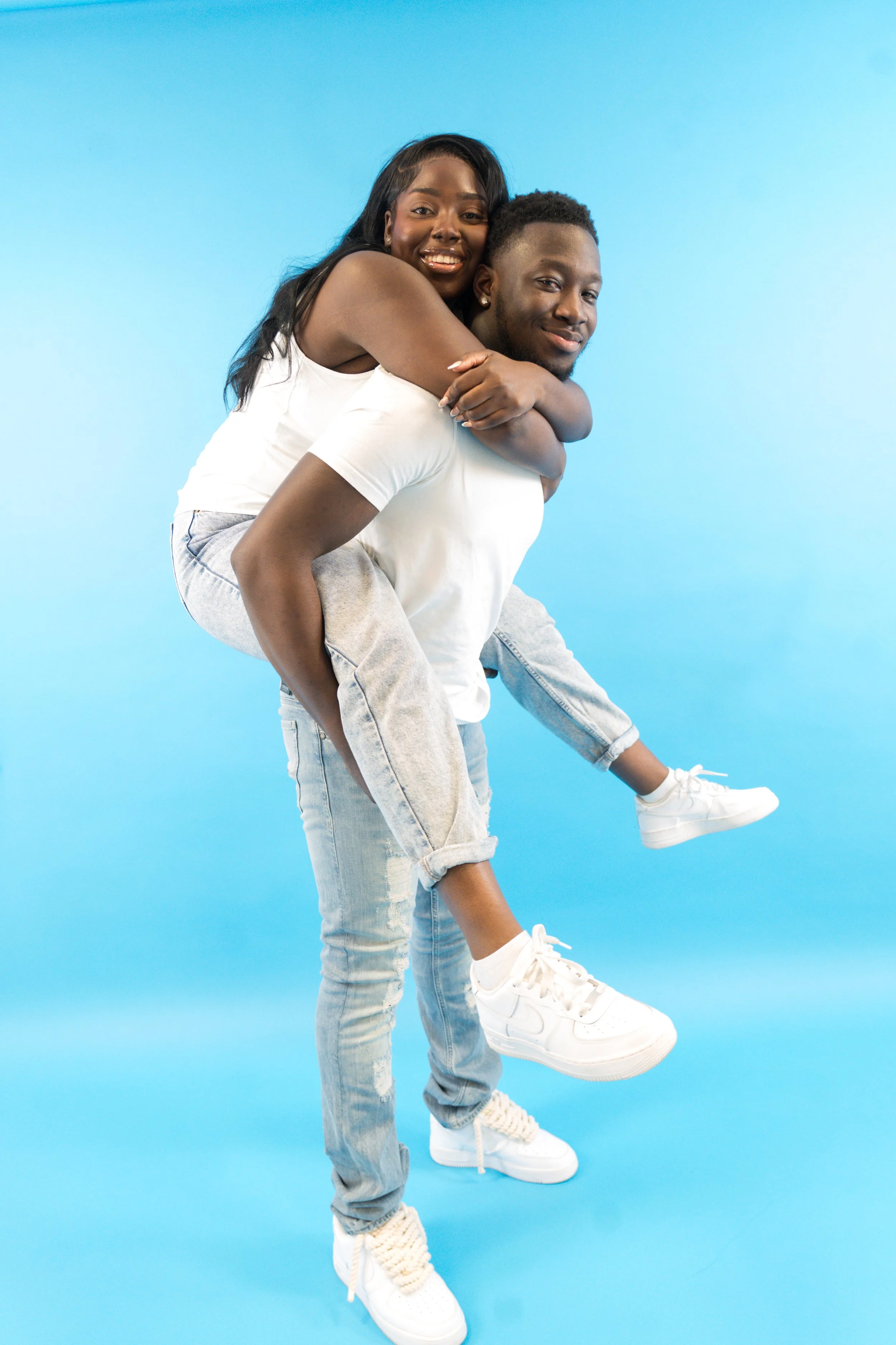 A joyful young woman is riding on the back of a young man, both smiling, against a light blue background.