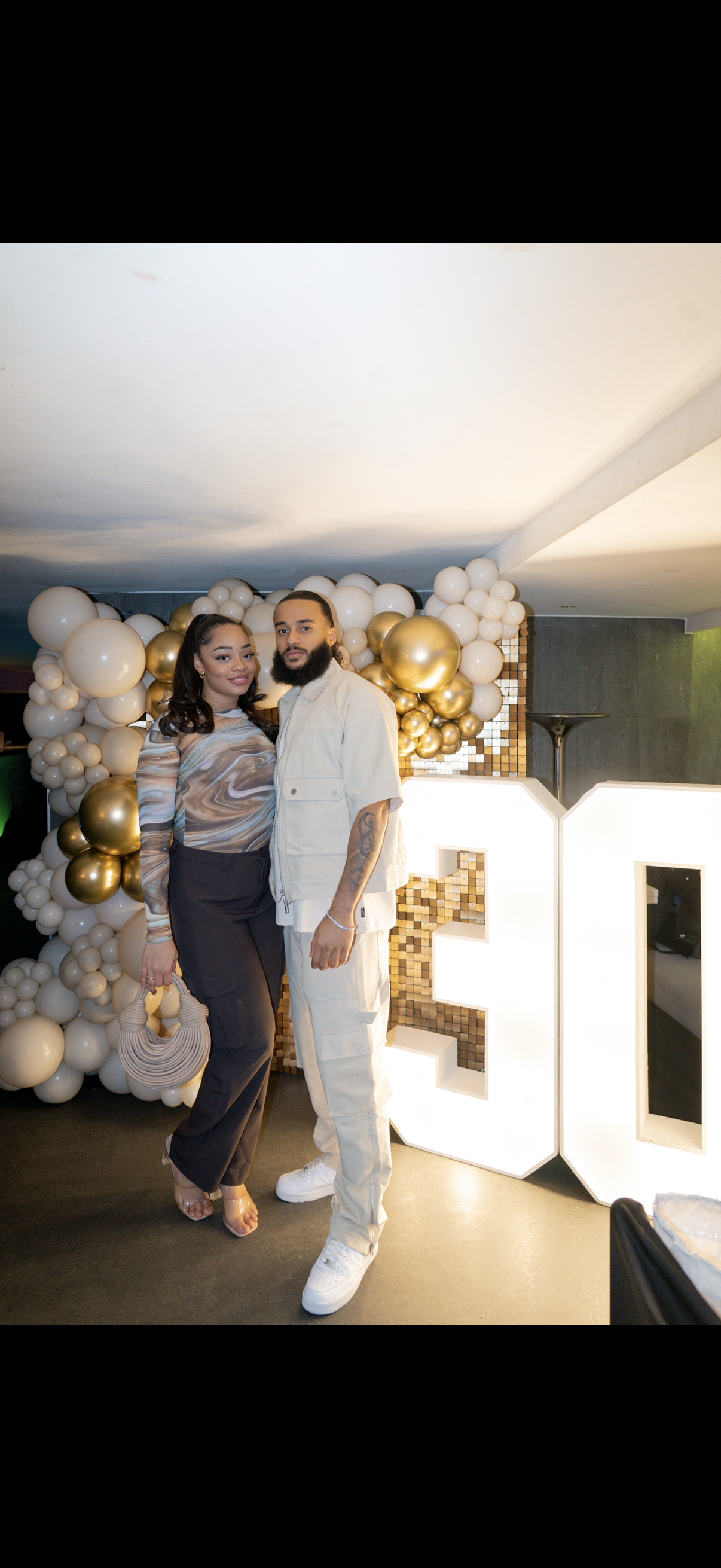 A couple standing closely together at a celebration, with a balloon arrangement made of white and gold balloons in the background, and the illuminated letters "E" and "O" beside them.