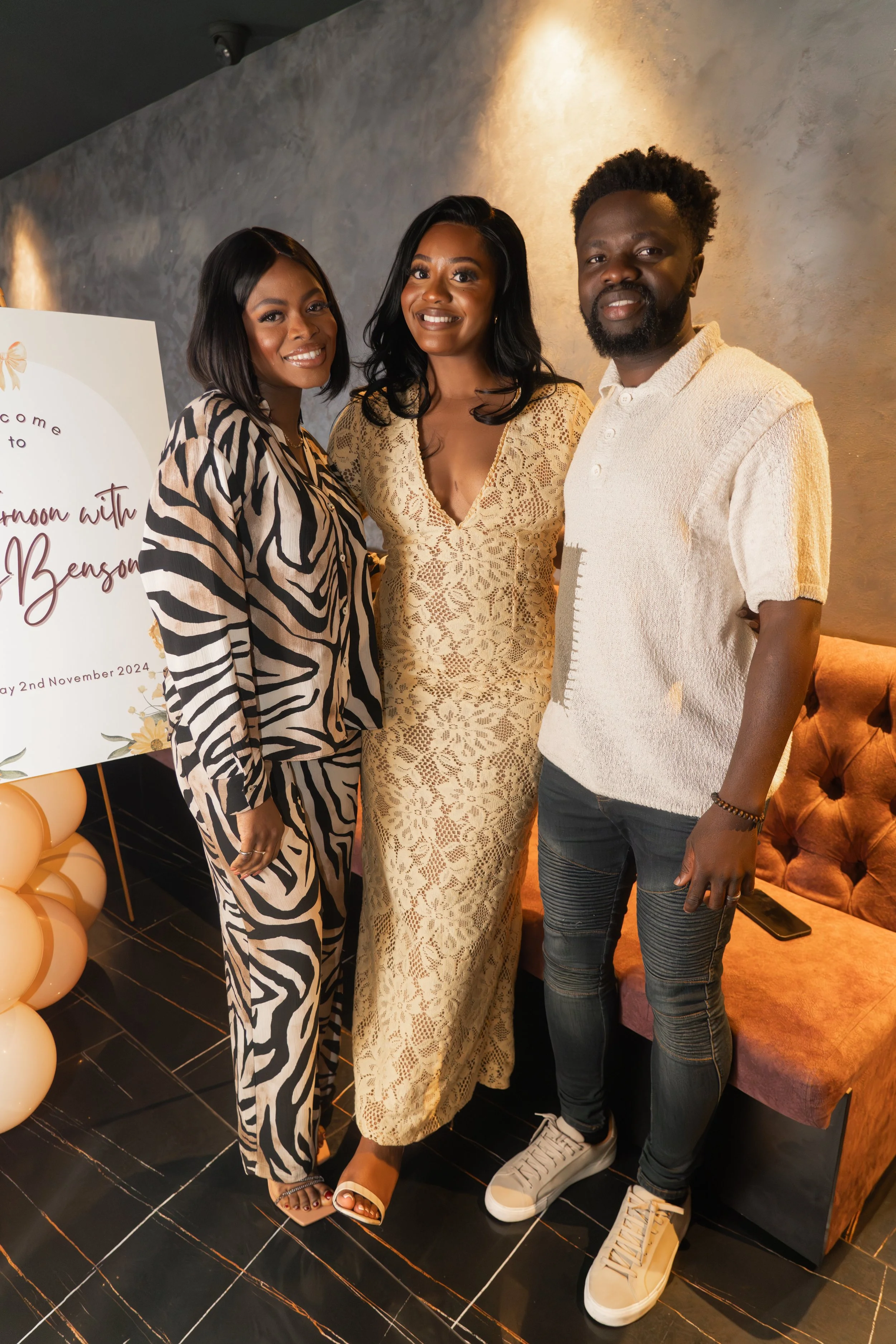Three people, two women and one man, posing together indoors at an event, with a sign about a baby shower in the background.