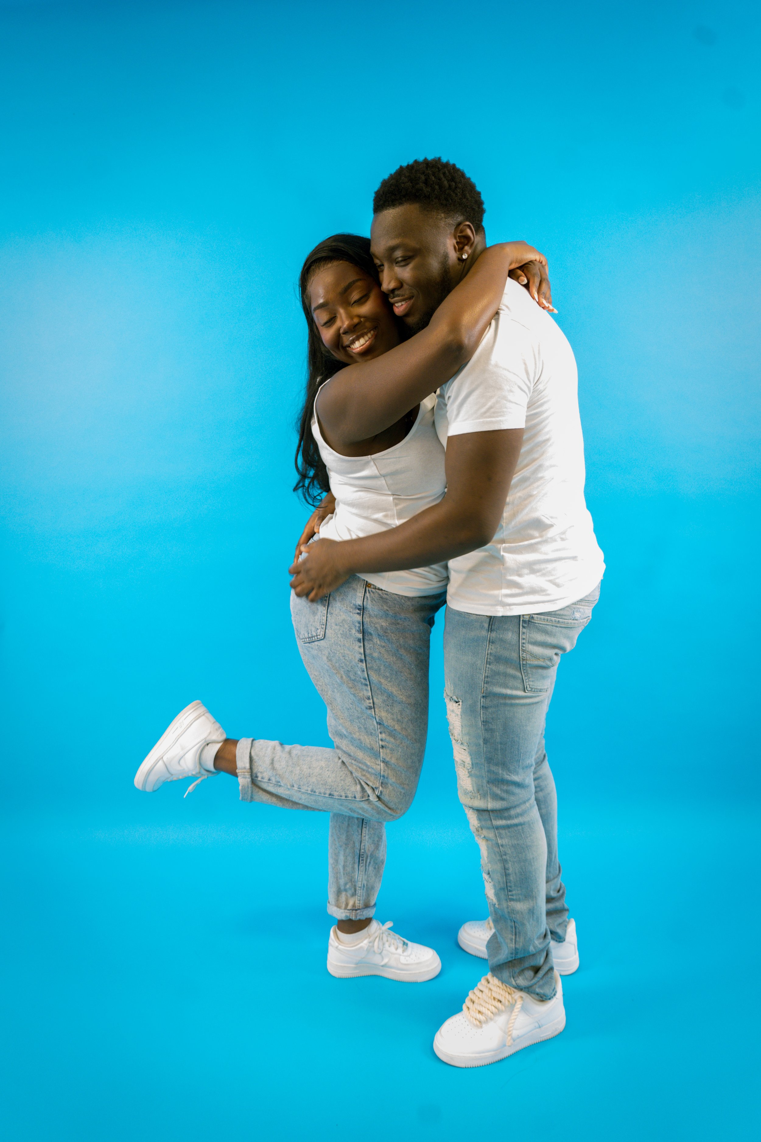 A young couple hugging and smiling against a blue background, both dressed in white t-shirts and light denim jeans.