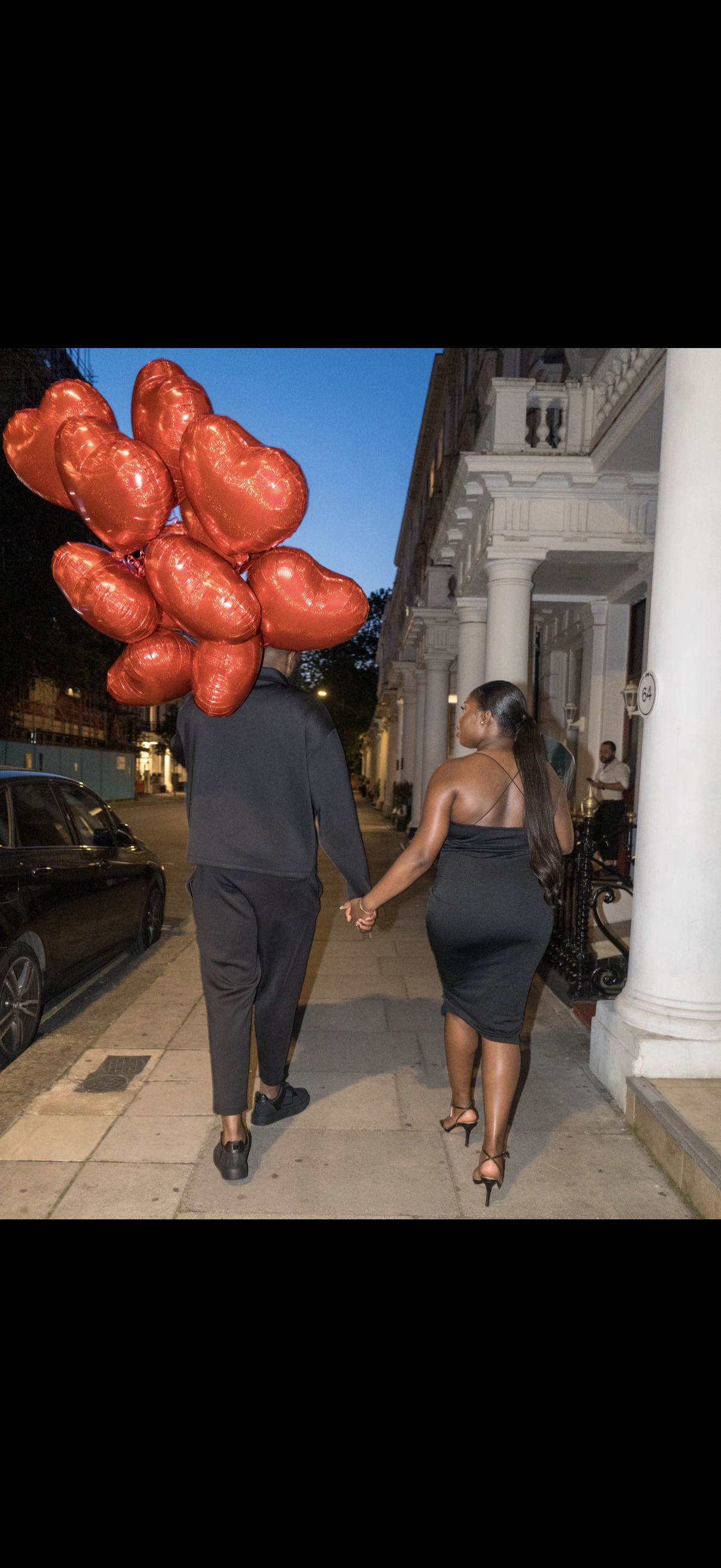 A couple walking hand in hand on a city sidewalk at dusk. The man is holding a large bunch of red heart-shaped balloons. The woman is wearing a black dress with high heels, and they are walking past a white, columned building.
