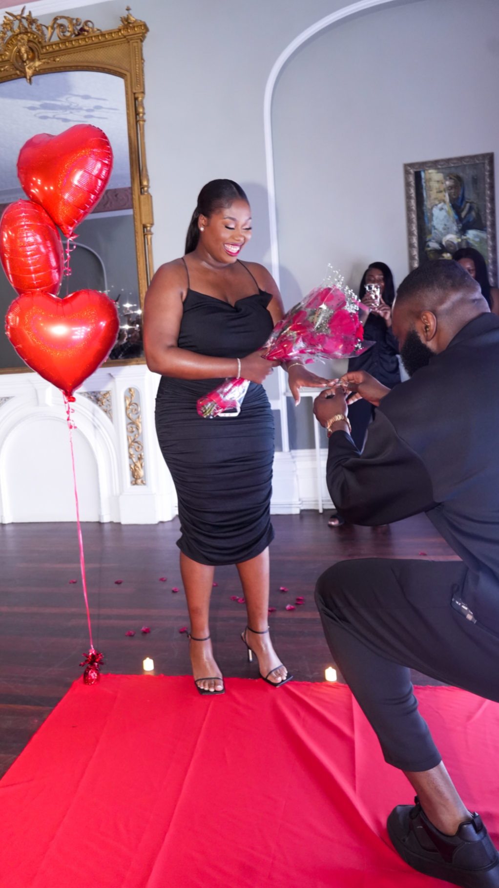 Woman in a black dress receiving a bouquet of pink roses from a man during a romantic proposal at an indoor venue decorated with red hearts and roses.